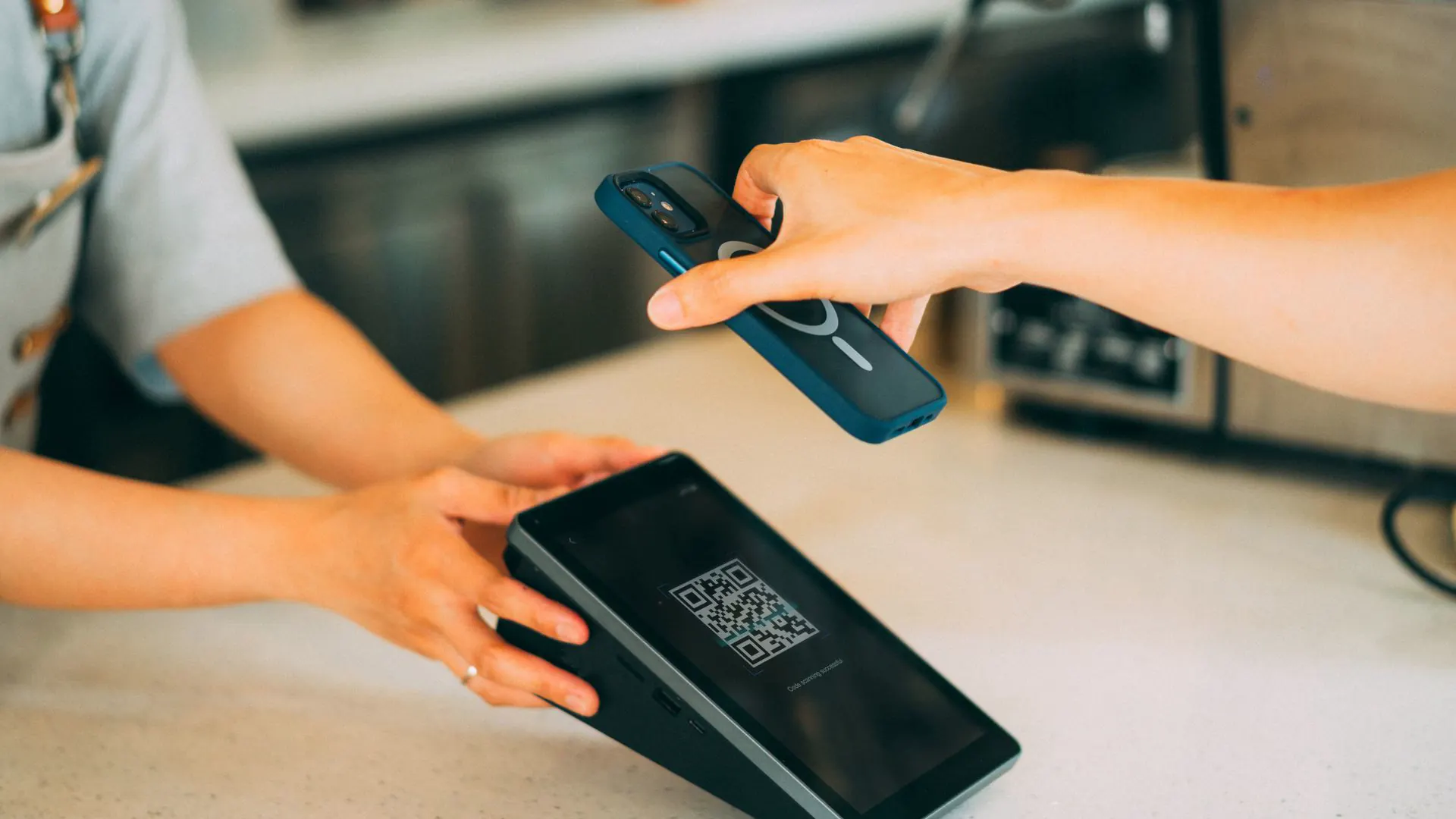 A customer holds a phone over a QR code payment terminal at a shop counter